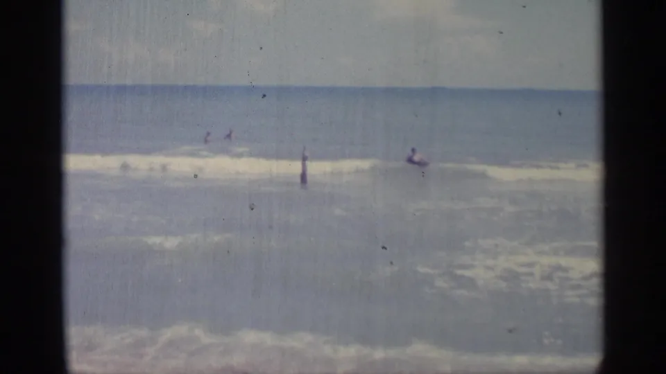 Wide 8mm frame from the Indiana beach scene showing swimmers dispersed in the surf under a pale sky.