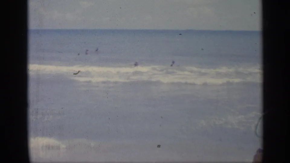 Soft 1959 Indiana 8mm frame with distant bathers in the surf and a broad waterline under bright daylight.