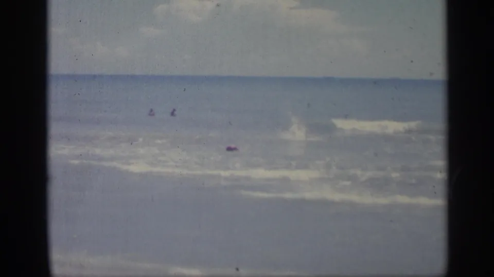 A 1959 home-movie view of beach surf shows small distant figures in the water and a breaking wave, with faded 8mm color.