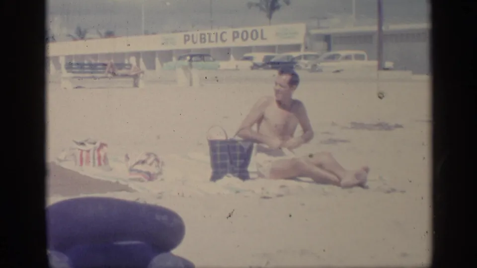 In this 1959 Indiana beach scene, a shirtless man sits on sand beside a bag while a public pool building and parked cars appear behind him.