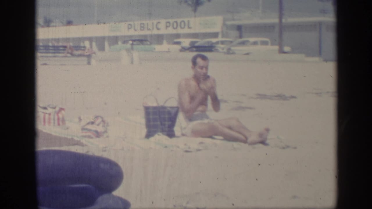 Man in swim trunks sits on sand near a public pool sign and parked cars.