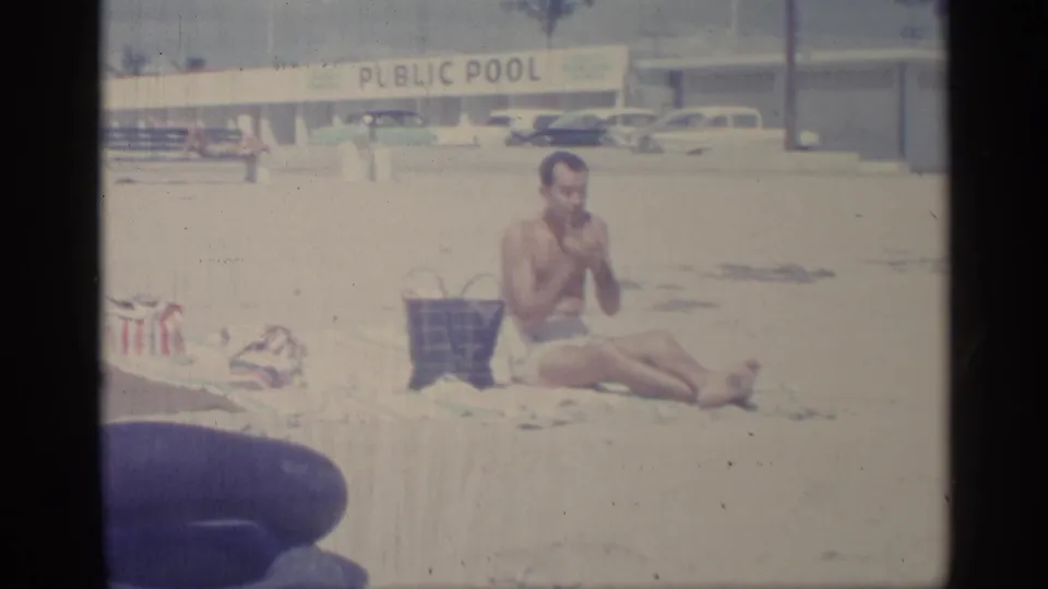 In 1959 Indiana home-movie footage, a man sits on a sandy beach near a public pool sign, with a plaid bag and cars behind him.