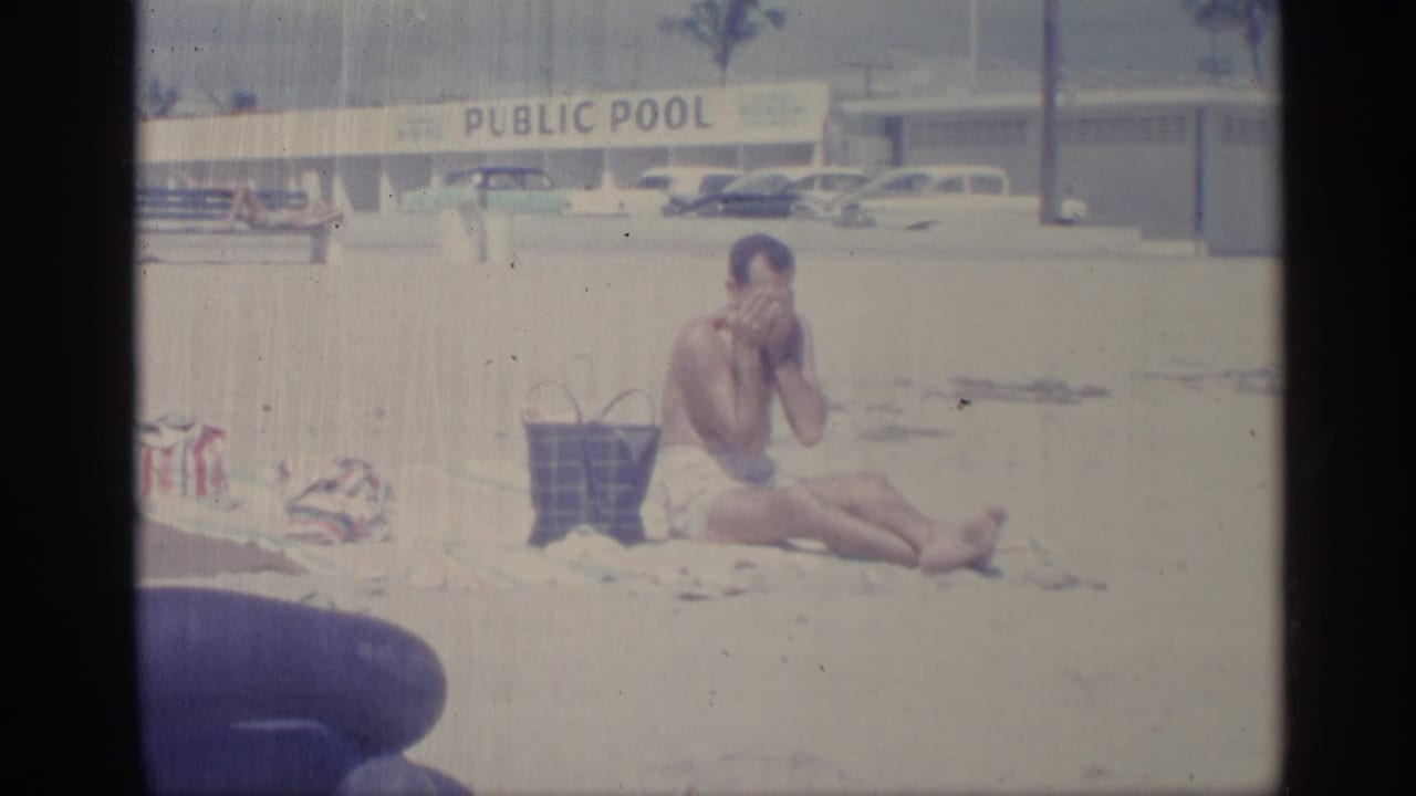 A seated man rubs lotion on his face near a public pool sign, with beach items in the foreground and parked cars in the background.