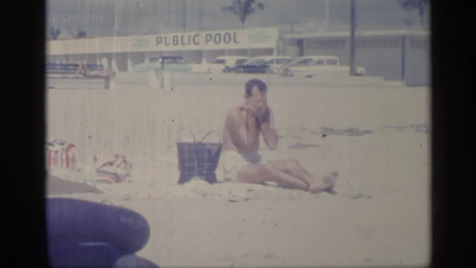 A seated man rubs lotion on his face near a public pool sign, with beach items in the foreground and parked cars in the background.