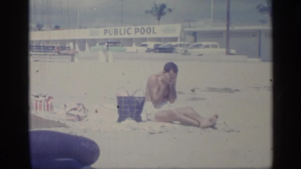 The man leans forward while rubbing lotion onto his face, with a public pool sign, parked cars, and beach belongings visible behind and beside him.