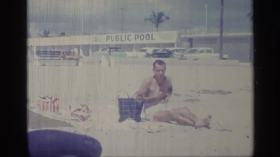 A man in swim trunks sits beside a plaid handbag on a sandy beach area, with a public pool sign and vintage cars in the background.
