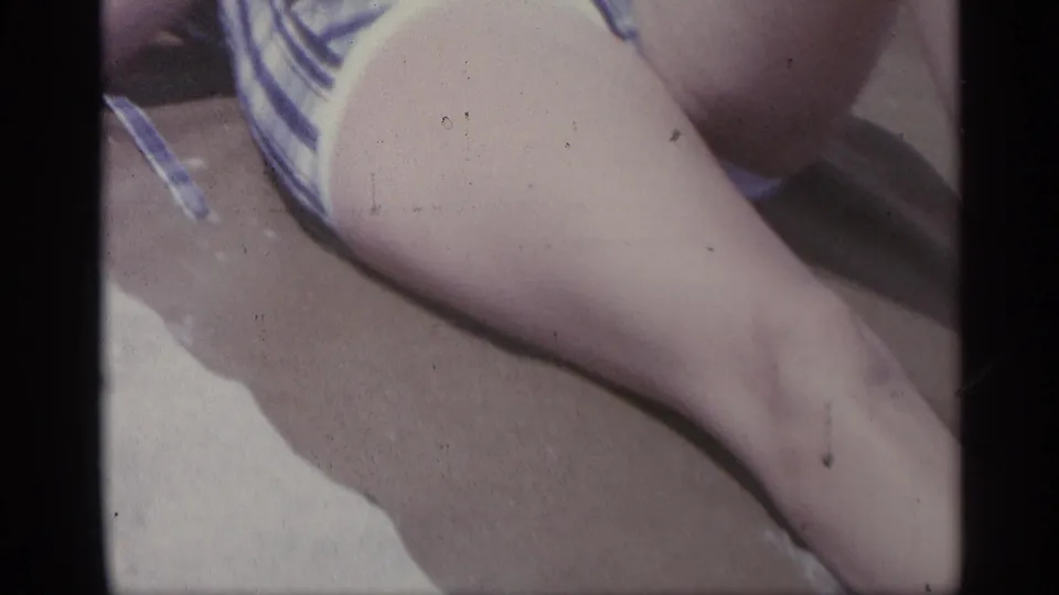 The home-movie frame focuses on the woman's legs and plaid swimsuit as she reclines on the sandy beach.