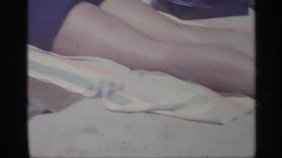 1959 Indiana home-movie frame with a close view of bare legs resting near a white cloth on a striped towel.