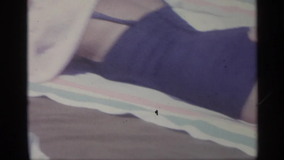 1959 Indiana 8mm frame showing a woman in a purple swimsuit reclining on a striped beach towel, with her face out of frame.