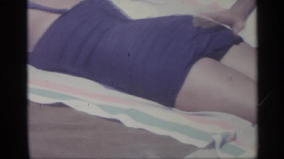 1959 Indiana home-movie frame showing a woman in a purple swimsuit lying sideways on a striped towel in soft, grainy color.
