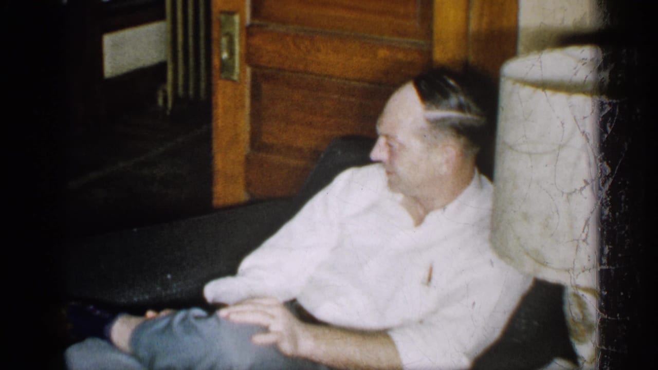 In a 1959 Michigan home-movie frame, an adult man sits on a dark couch in a dim interior with wood doors behind him.