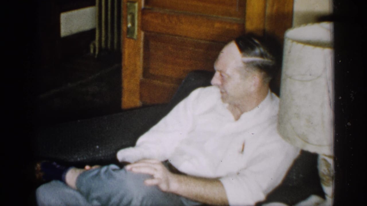 The man remains seated on the couch, turned slightly to one side, with a wooden doorway and pale lamp or column nearby.