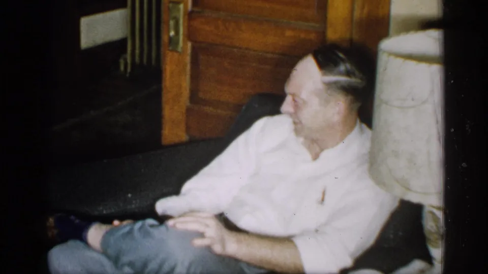 The man remains seated on the couch, turned slightly to one side, with a wooden doorway and pale lamp or column nearby.