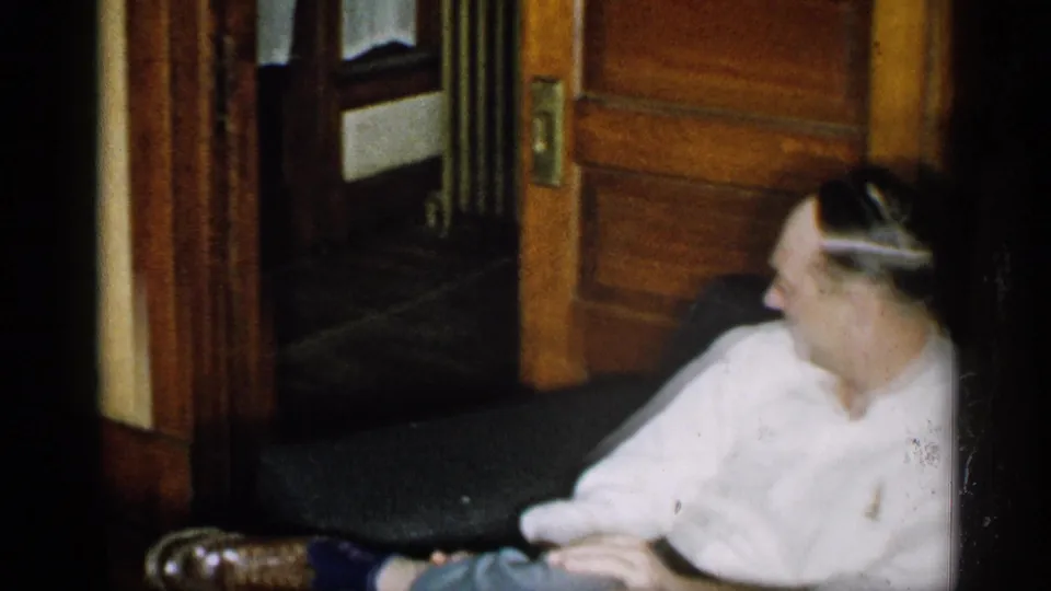 A soft 8mm frame shows the man partly cropped on the couch, with wood trim, a doorway, and dim room details in the background.