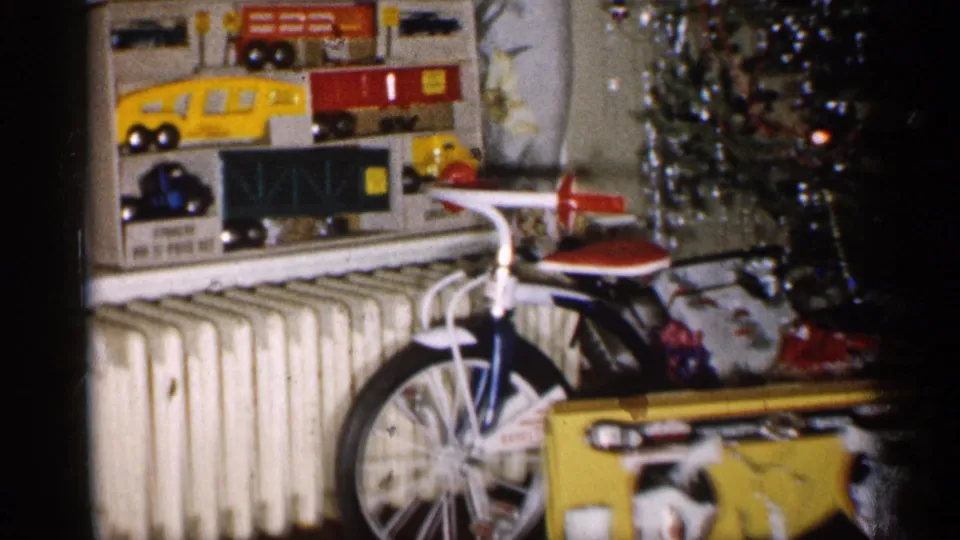 1959 Michigan home interior with a toy vehicle display, small bicycle, radiator, tinsel Christmas tree, and a large boxed gift in view.