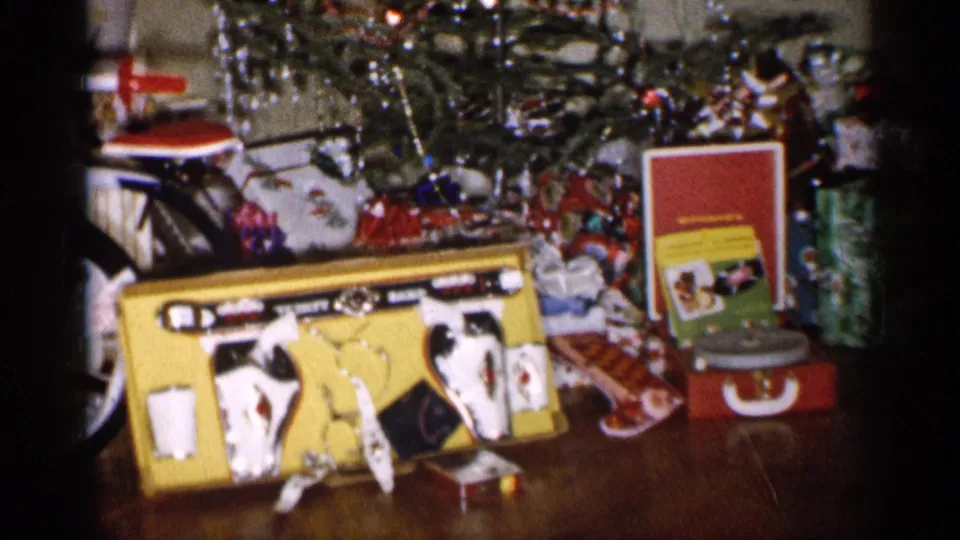 1959 Michigan home-movie frame showing a holiday gift display with bicycle, toy boxes, wrapped presents, tinsel, and a small tree stand.