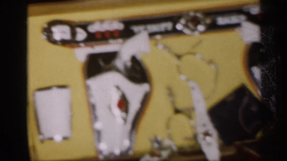 1959 Michigan home-movie close-up of toy pistols, a holster belt, and accessories on a yellow backing board.
