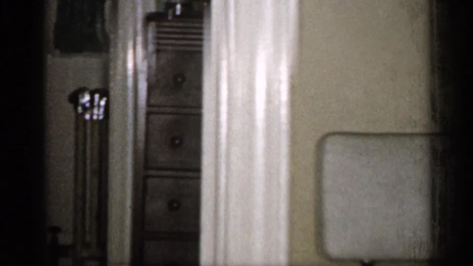 A home interior view shows a doorway, dresser drawers, radiator, and the top of a chair back in soft 8mm focus.