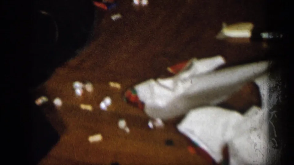 Low-angle 8mm view of Christmas wrapping debris, a white cloth or garment, and scattered small items on a wooden floor in Michigan.