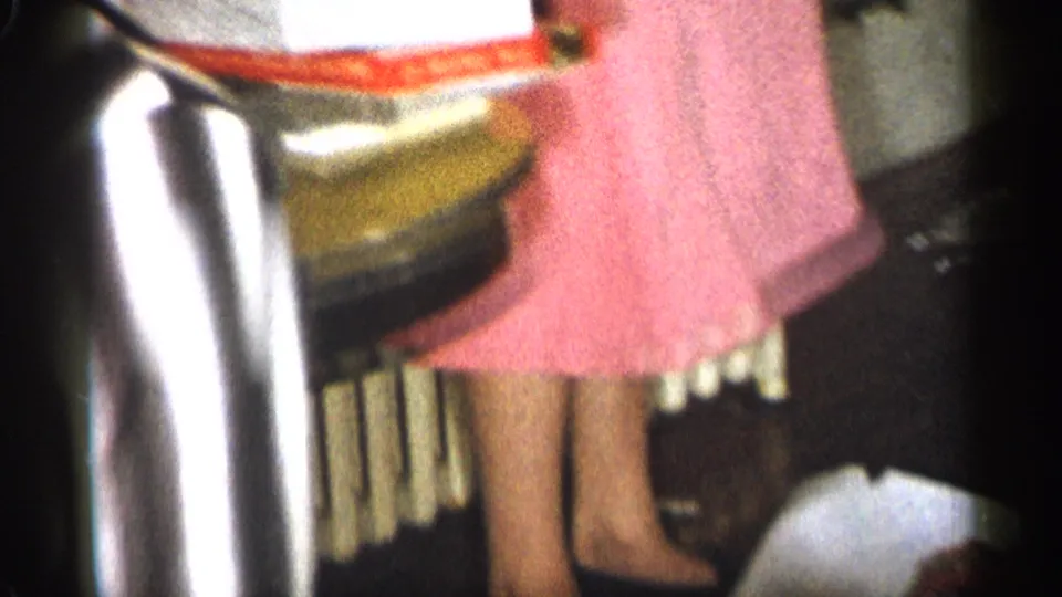 1959 Michigan home-movie frame showing party guests cropped at the waist, with a woman in a pink dress beside a radiator.