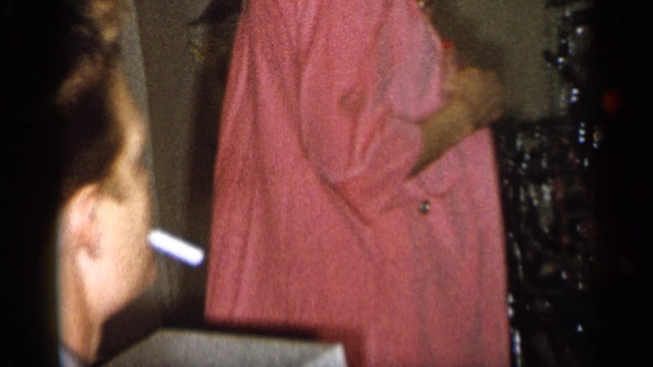 1959 Michigan interior party frame with a woman in a pink dress holding a cigarette holder near a seated person's head.