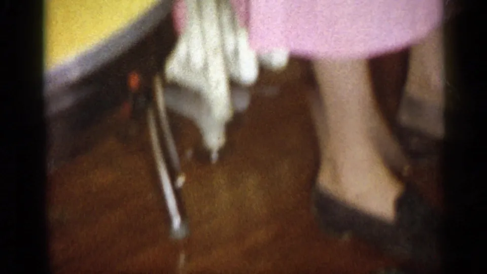 1959 Michigan home-movie frame showing a woman in a pink dress and dark heels standing beside a chair in a private room.