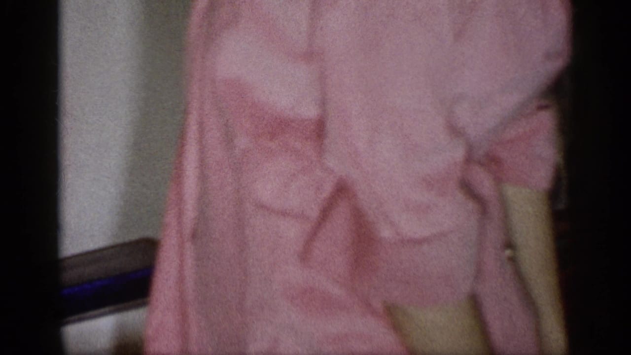 Cropped 1959 Michigan home-movie frame of a person in a pink dress inside a private room during a holiday gathering.