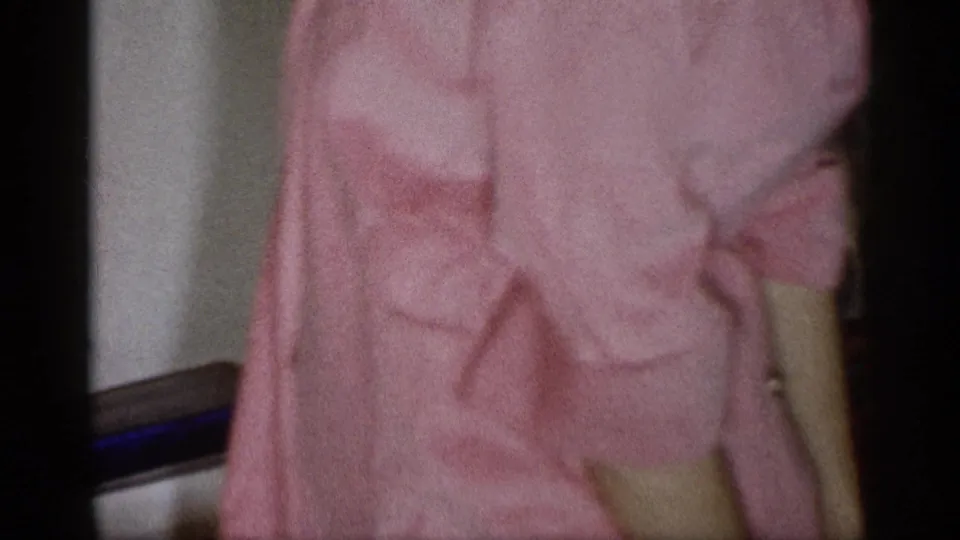 Cropped 1959 Michigan home-movie frame of a person in a pink dress inside a private room during a holiday gathering.