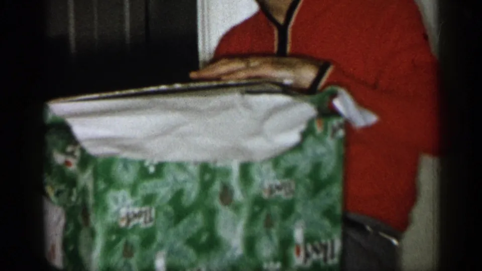 1959 Michigan home-movie frame showing hands opening a green holiday gift box with a red-shirted person in the background.