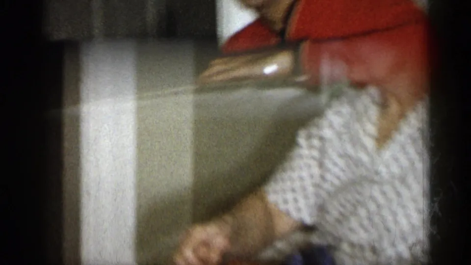 1959 Michigan home-movie frame of people handling a gift box indoors, with a checkered sleeve and red shirt visible.
