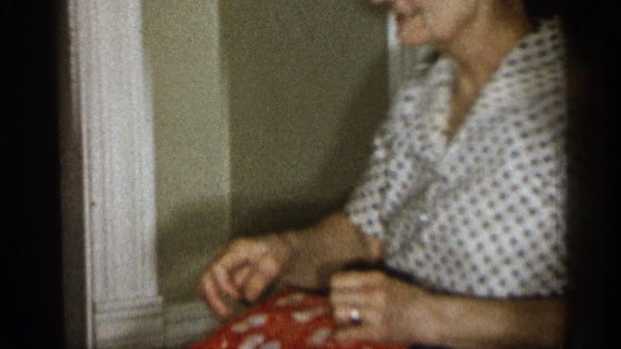 Elderly woman in glasses sits indoors with hands near a red patterned cloth.
