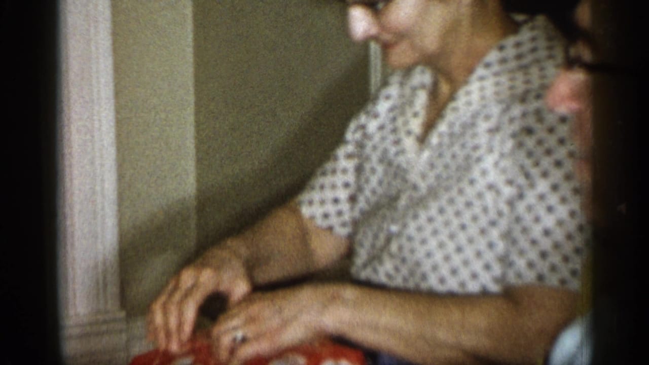 The woman looks down toward her hands as she handles a red cloth in a private Michigan interior, consistent with 1959 home-movie footage.