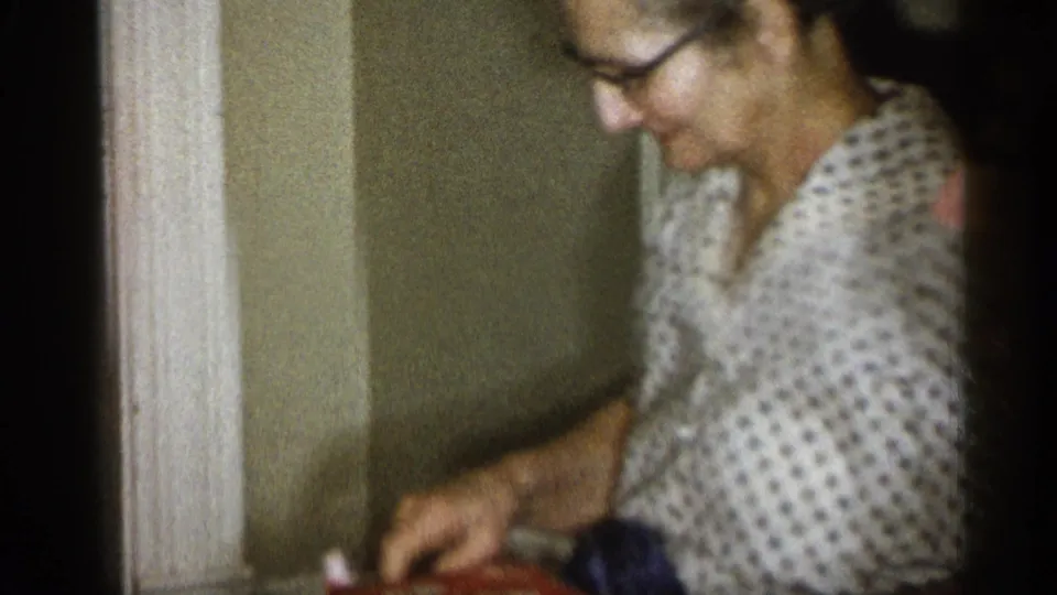 A seated older woman leans forward in a softly focused 8mm frame, reaching toward a red patterned cloth inside a Michigan home.
