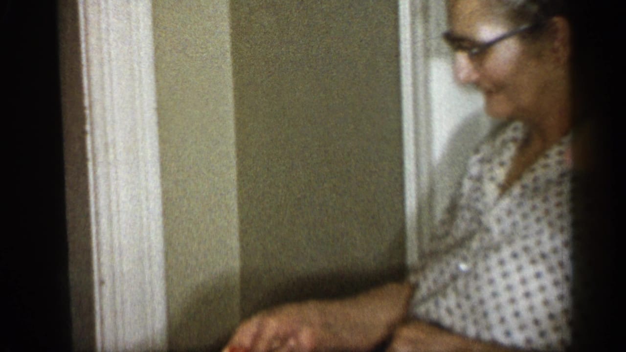 Woman in glasses sits indoors near a doorway while handling something out of frame.