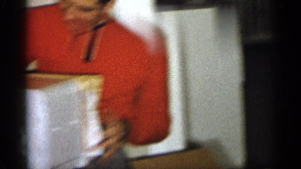 Man in a red shirt stands indoors holding or opening a colorful box.