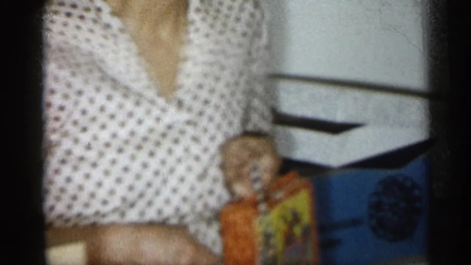 Close 8mm frame of the woman in a polka-dot blouse holding a colorful box, with her head partly cropped and the private interior behind her.