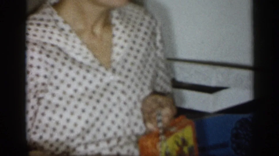 The woman continues holding the colorful box in a softly focused 1959 Michigan home-movie frame, with the interior wall and trim visible behind her.