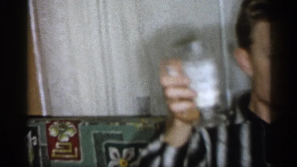 Adult man in a striped shirt holds up a glass inside a Michigan home interior in 1959 8mm footage.