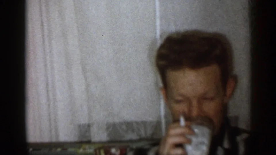 Adult man seated on a couch drinks from a glass inside a Michigan home interior in 1959 8mm footage.