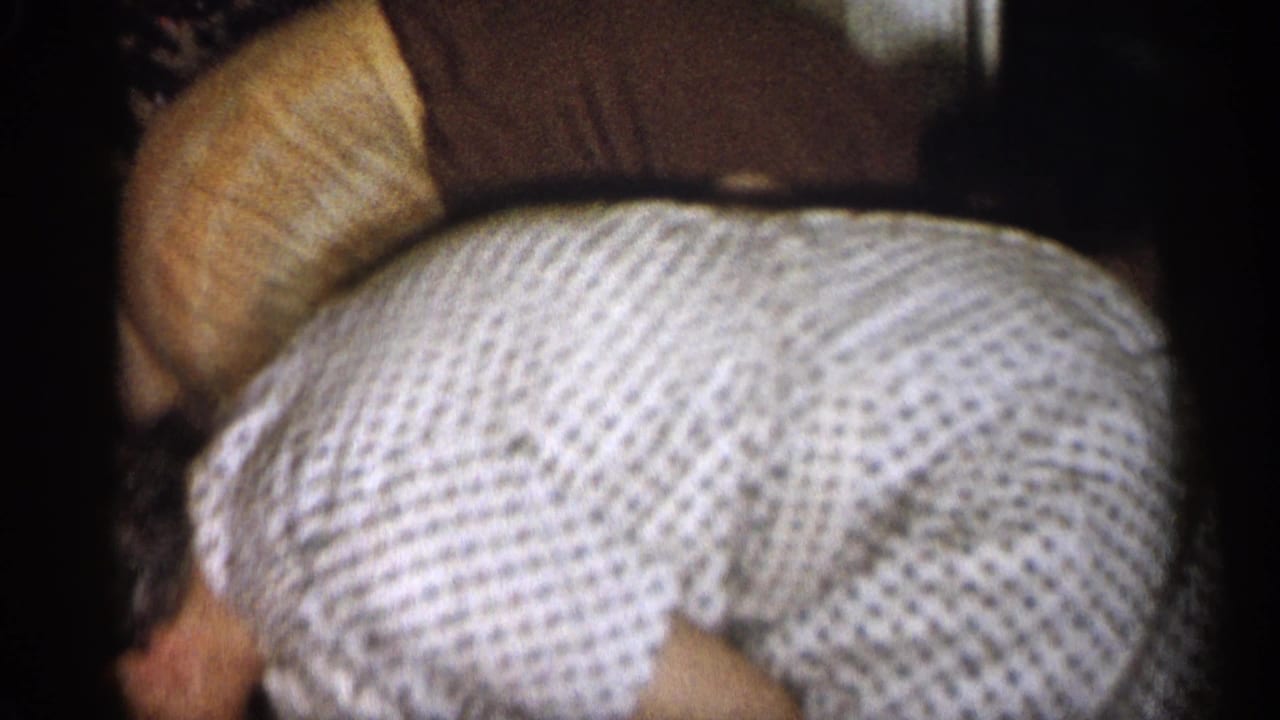 1959 Michigan home-movie frame showing two adult women bent over beside upholstered furniture in a private interior.
