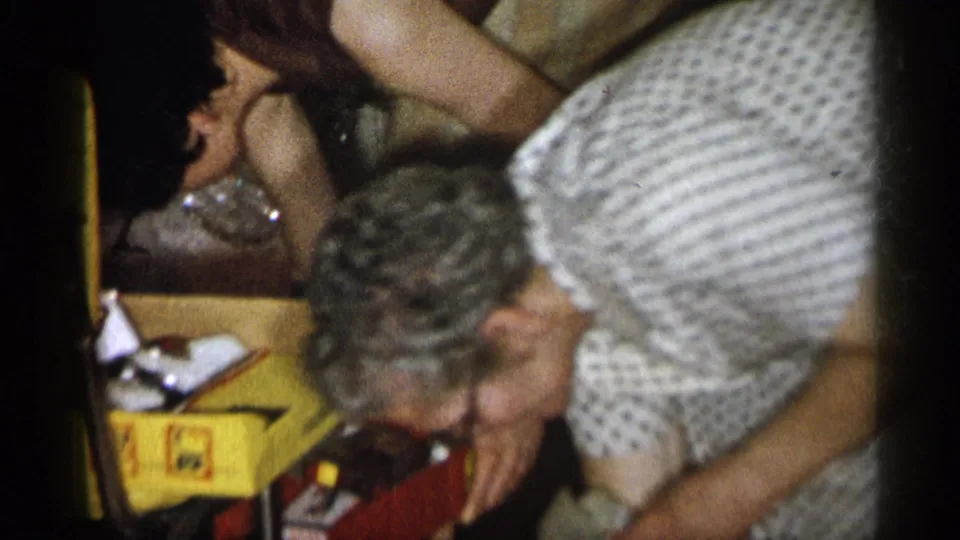 1959 Michigan 8mm frame of a woman in a patterned dress reaching near a red box, yellow box, and loose papers indoors.