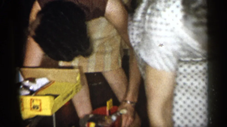 1959 Michigan home interior frame with two adult women bending over boxes, papers, and small household items on the floor.