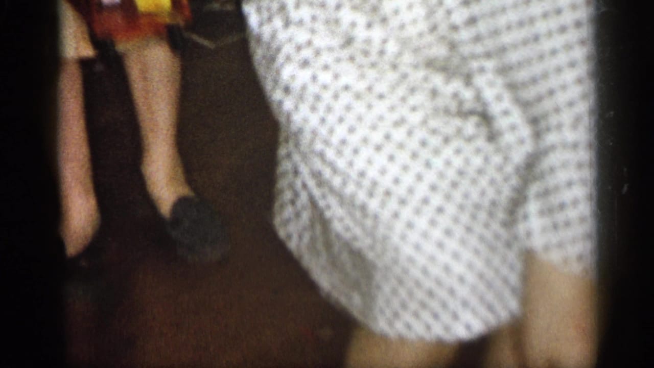 Adult in a white polka-dot dress moves past a bed or low furniture inside a dim room.