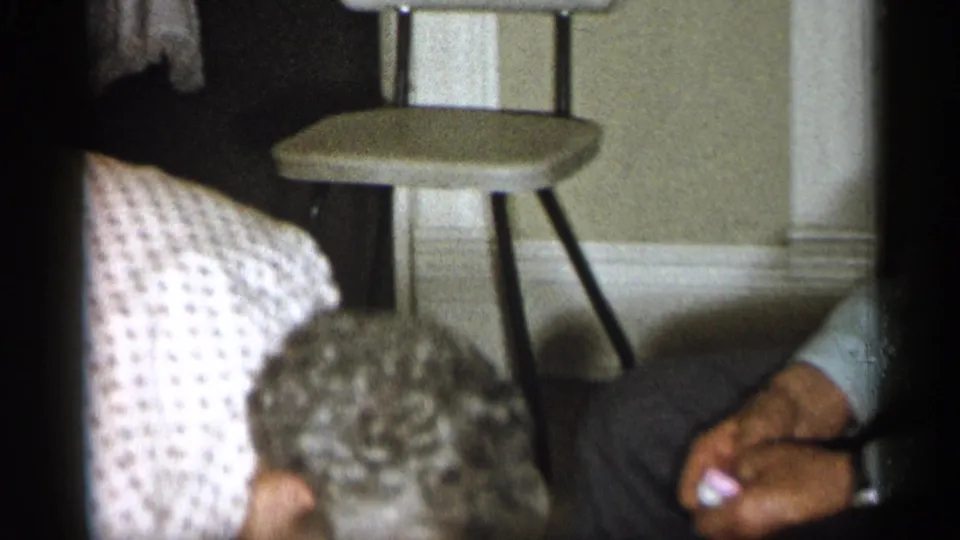 1959 Michigan home-movie frame with a person in a striped shirt leaning near a small stool and furniture in a private interior.