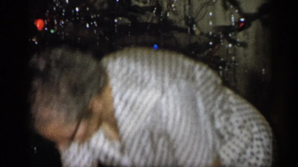 1959 Michigan home-movie frame showing an adult in a patterned dress bending in front of a decorated Christmas tree indoors.