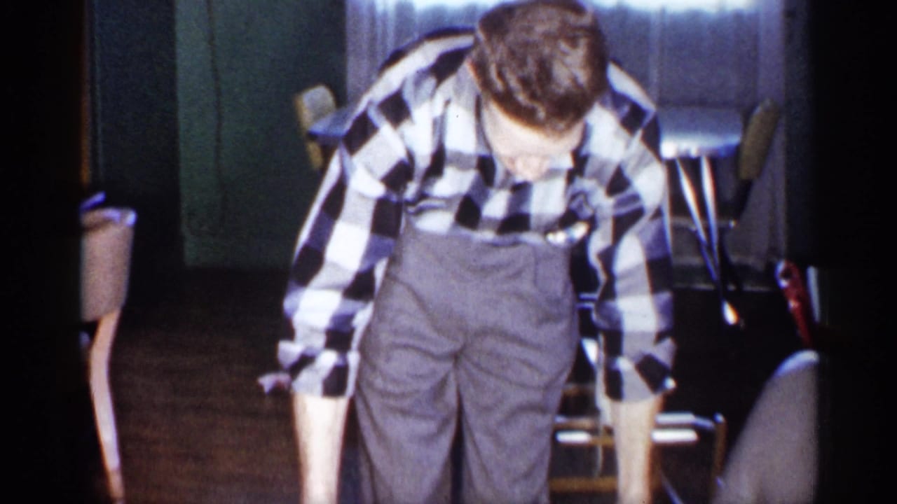 In a 1959 Michigan home interior, a man in a black-and-white checked shirt and grey trousers bends toward an unseen box.