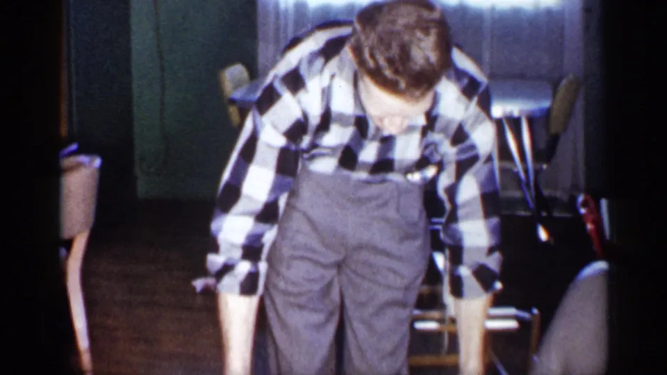 In a 1959 Michigan home interior, a man in a black-and-white checked shirt and grey trousers bends toward an unseen box.