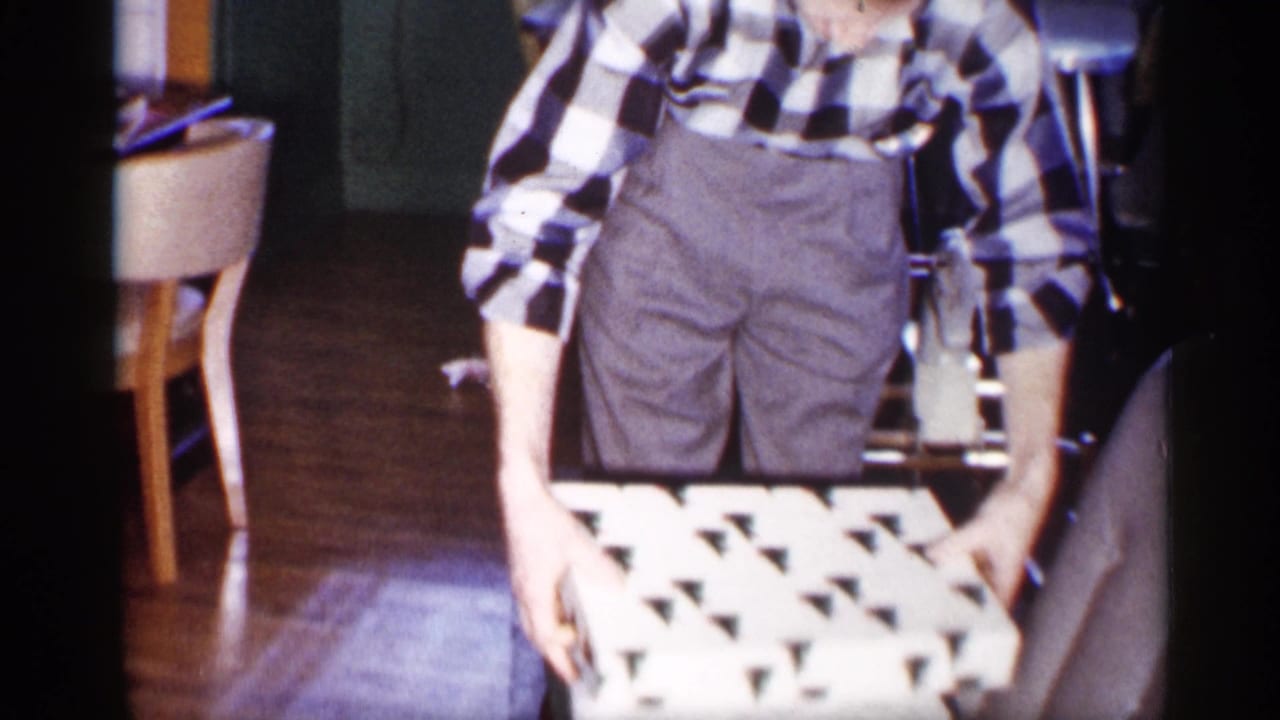 The man stands indoors holding a green-and-white patterned gift box, with chairs and wooden flooring behind him.