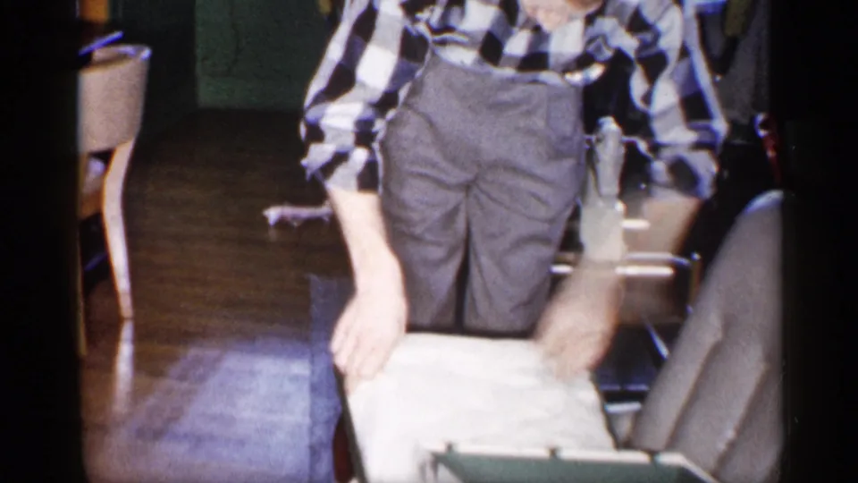 The man bends down over a pale box lid or wrapping, continuing the gift-opening action inside the home.