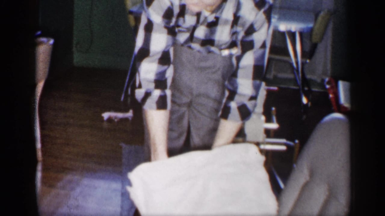 Man in checkered shirt stands indoors holding a white cloth near folding chairs.